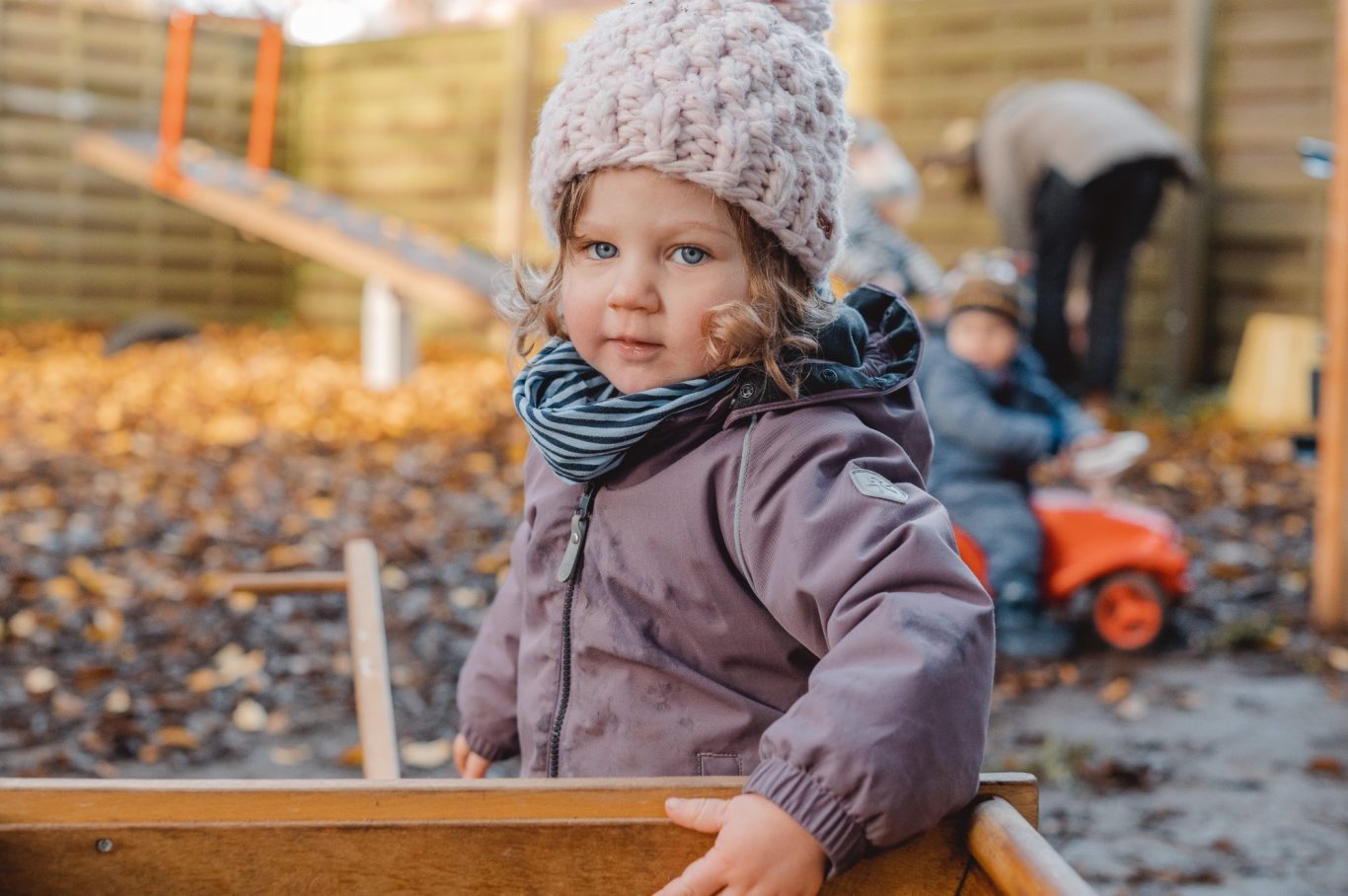 Kleines Mädchen in warmer Jacke und Mütze, spielt im Laub mit anderen Kindern im Hintergrund.