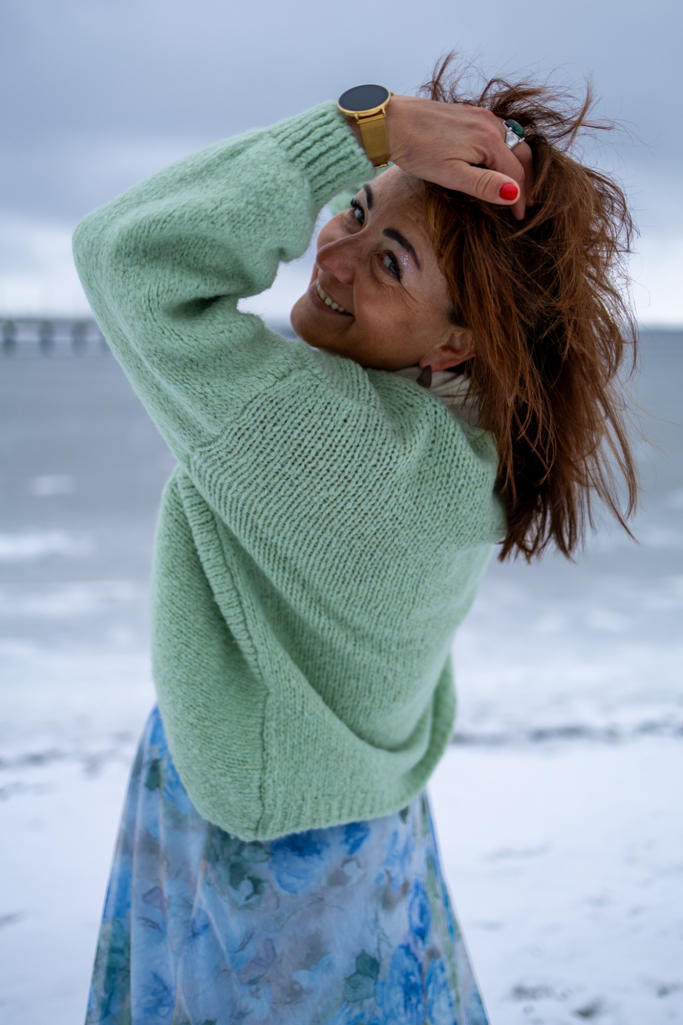 Frau mit lockigem braunem Haar, trägt einen grünen Pullover und steht am Strand.
