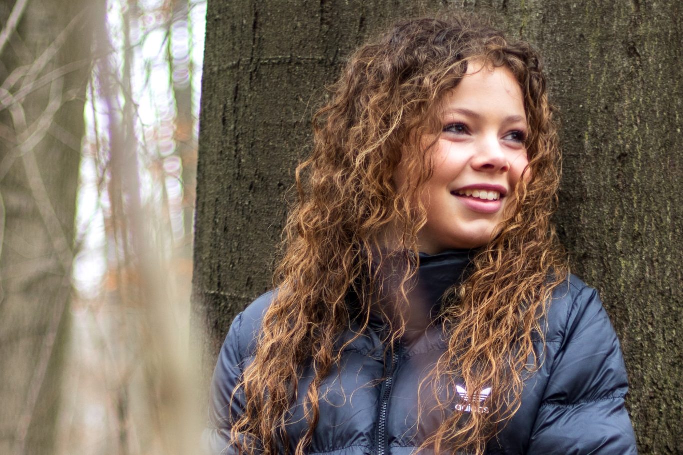 Junge Frau mit lockigem, braunem Haar lächelt an einem Baum im Wald.
